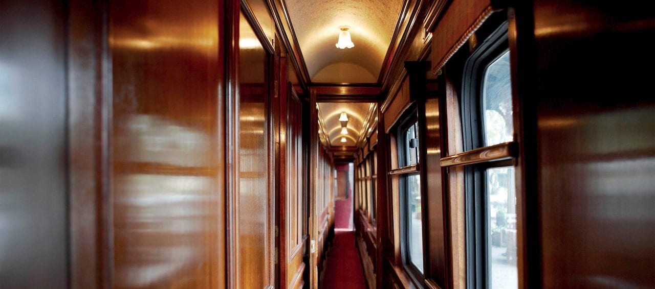 Interior view of the Belmond Royal Scotsman train corridor, featuring polished wood paneling, red carpet, and exterior views through windows.