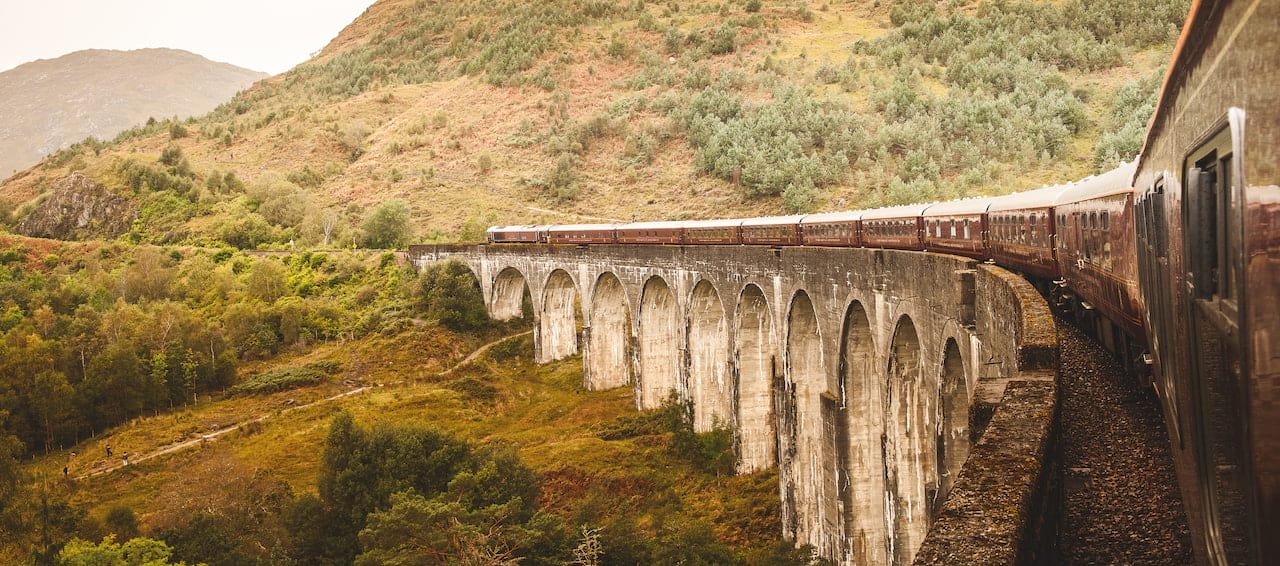 Belmond Royal Scotsman train traverses the Glenfinnan Viaduct, Scotland, on the Clans, Castles and Isles tour. Lush hillside backdrop.
