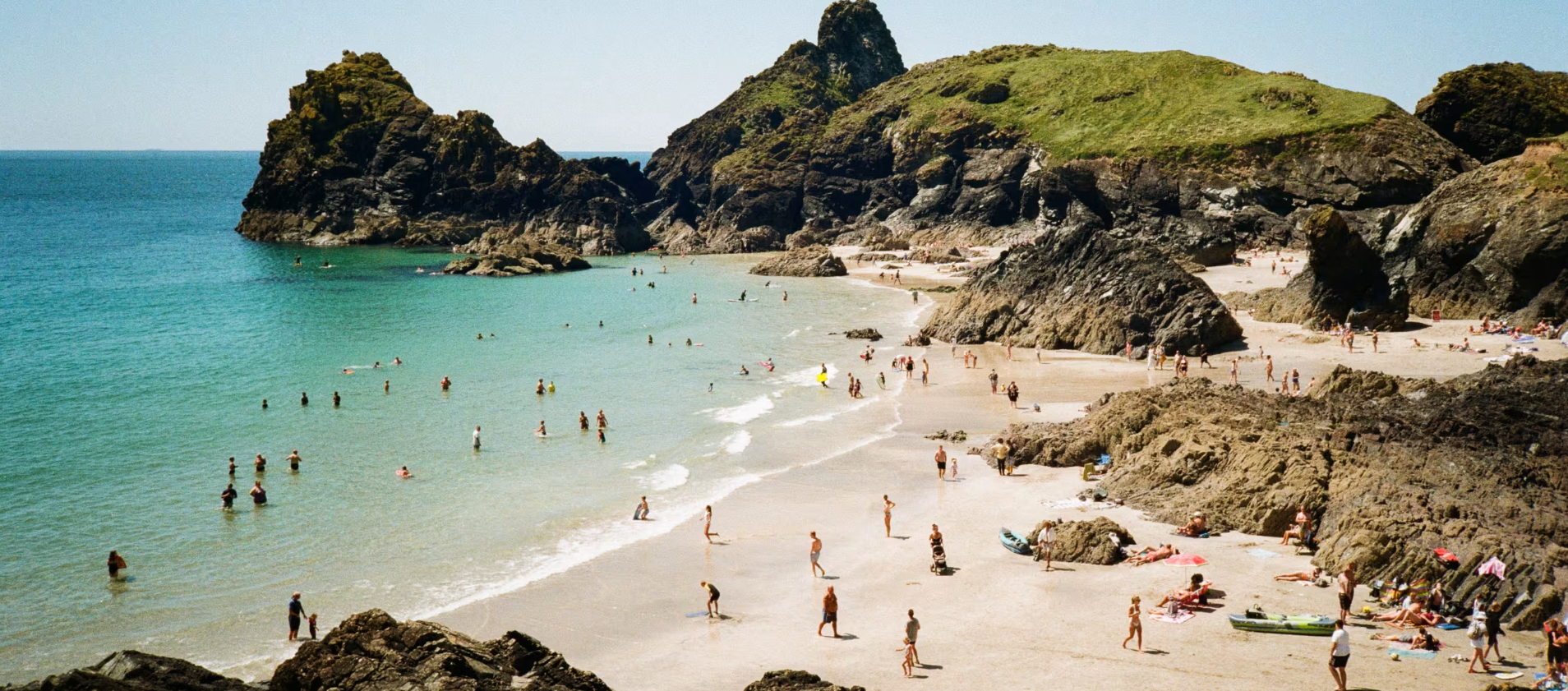Crowded beach in Cornwall, UK, with rocky cliffs and turquoise water. People swimming, sunbathing, part of Britannic Explorer tour.