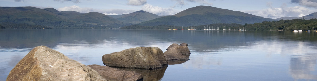 Still waters of Loch Lomond, Scotland, with mountains in the distance. Sailboats dot the horizon. Rocks in foreground. Belmond tour.