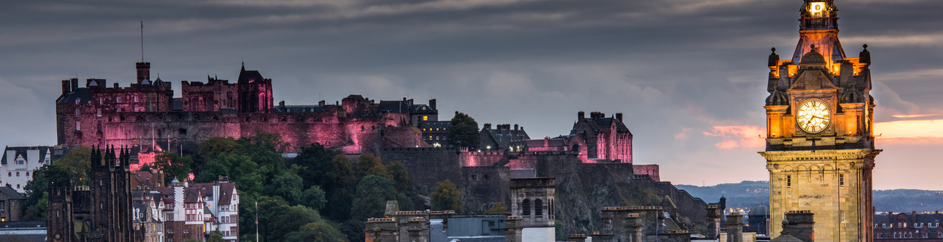 Edinburgh Castle illuminated pink at dusk, seen on the Belmond Royal Scotsman tour, with a lit clock tower in the foreground.